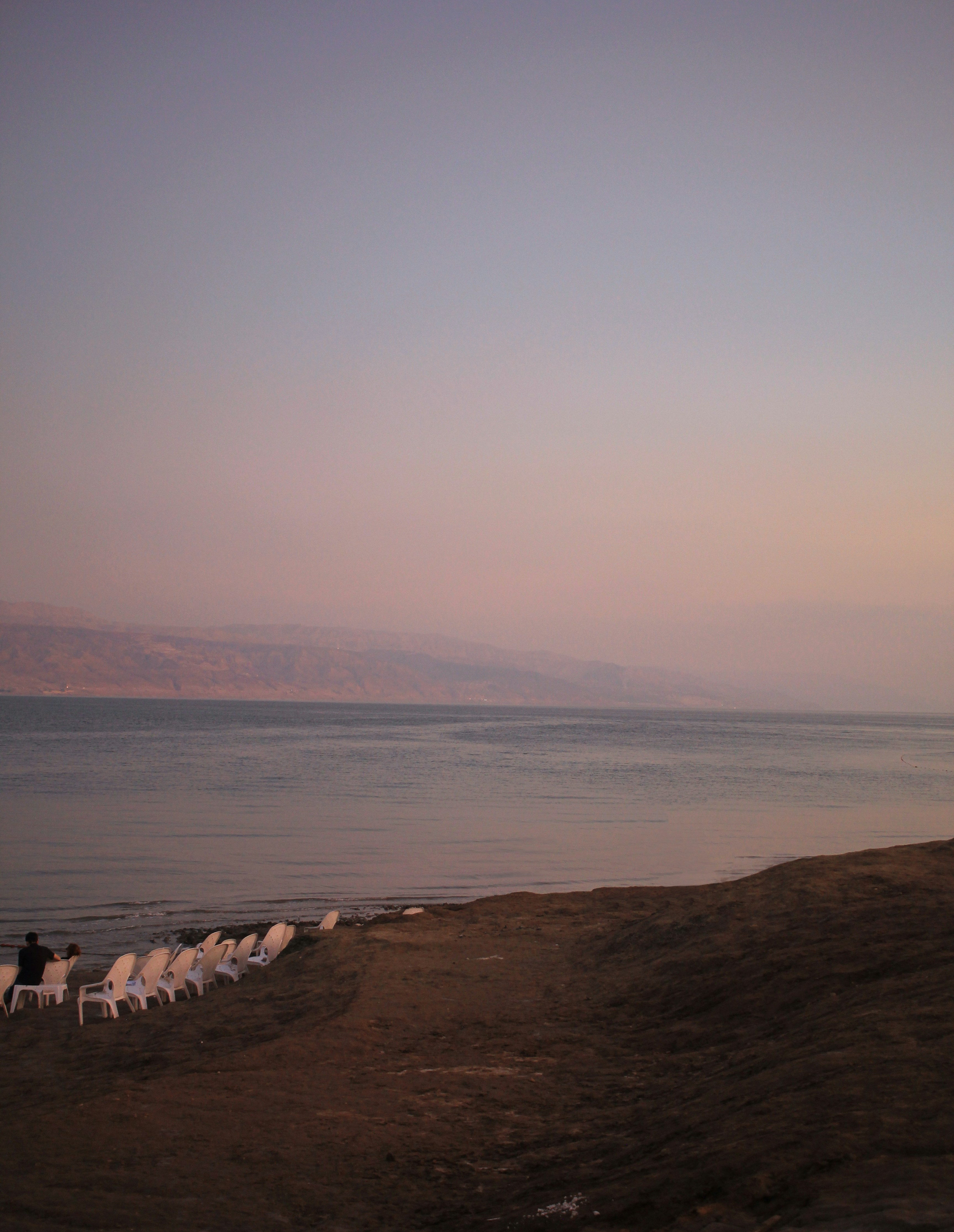 Dead Sea shoreline at dusk from the Jordanian side, with white plastic chairs and distant hills