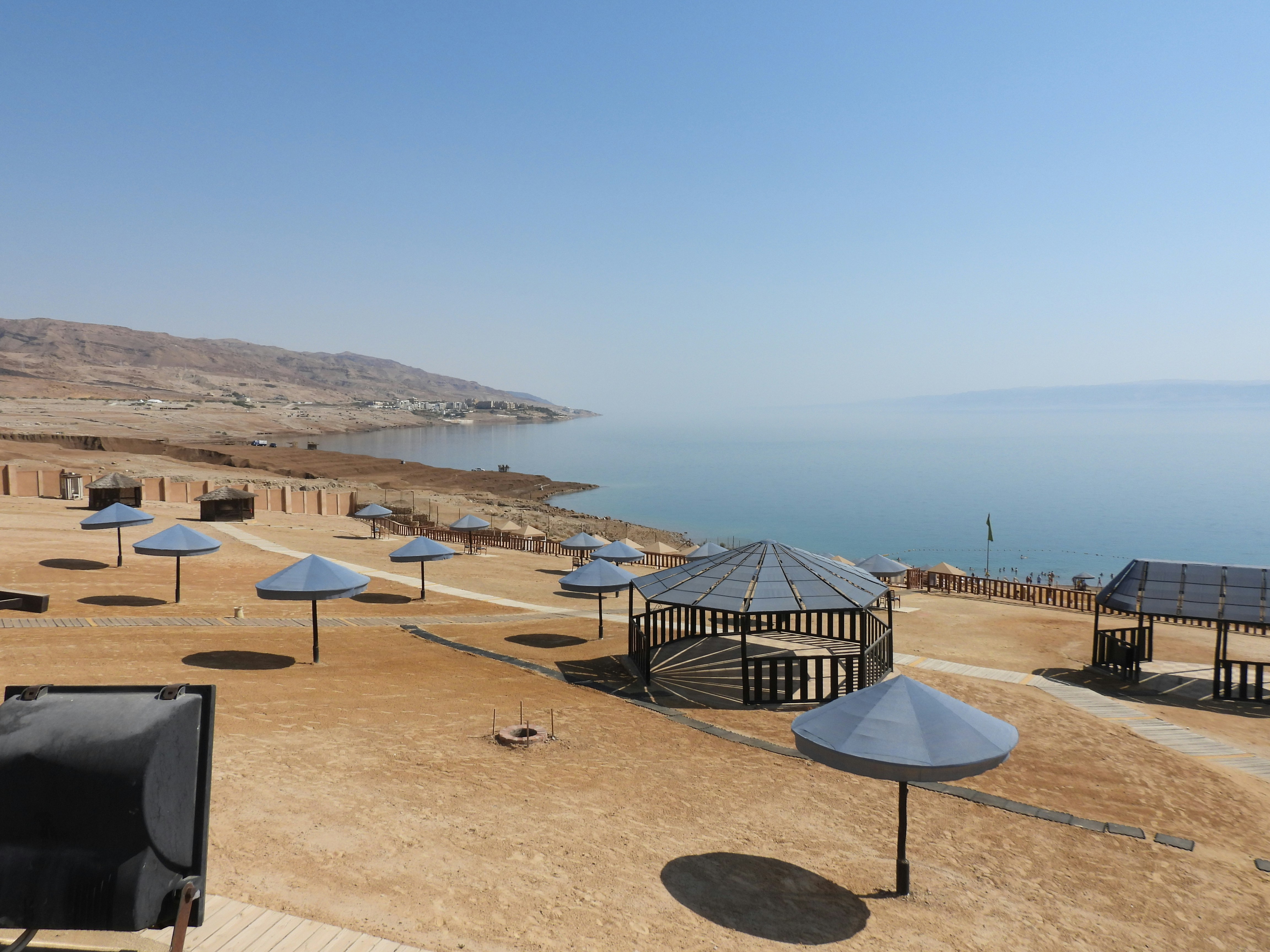Beach umbrellas and gazebos overlooking the calm Dead Sea shoreline with the Judean hills in the background