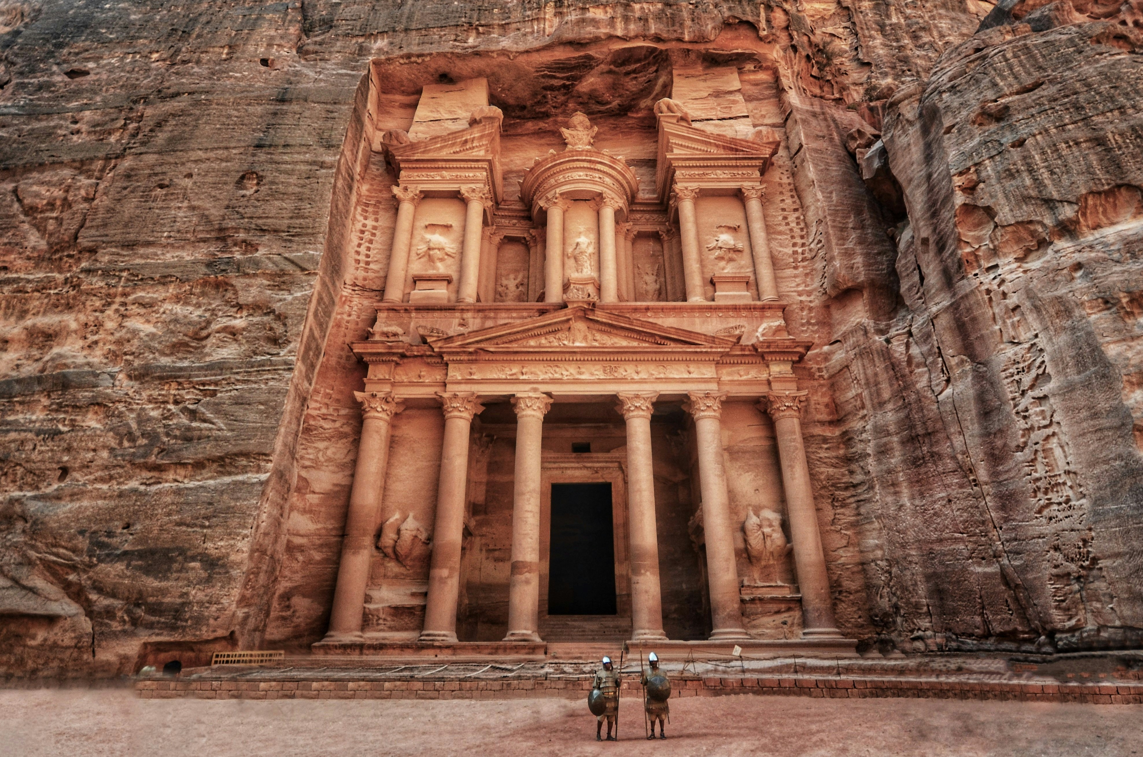 Two visitors standing in front of the magnificent Treasury facade carved into the rose-red sandstone cliffs at Petra, Jordan